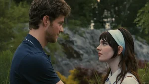 A man and woman stand facing each other outdoors, with greenery and rocks in the background. She wears a headband; both appear focused.