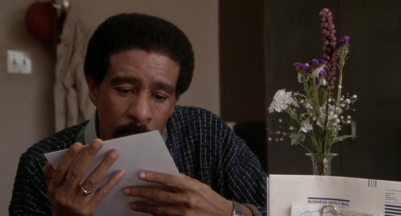 A man with short curly hair sits at a desk, reading a piece of paper, with a vase of flowers and mail in the foreground.