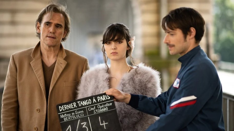 Three people stand together; one holds a movie clapperboard labeled "Dernier Tango à Paris." They appear to be on a film set under an arched structure.