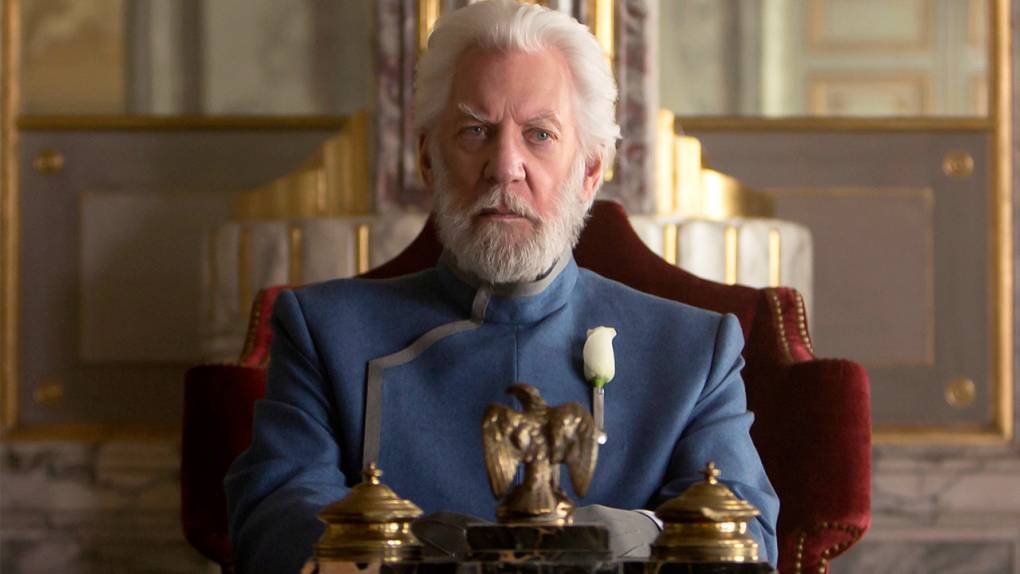 Older man with white hair and beard, wearing a formal blue suit, sits on a grand chair in an ornate room. A white rose is pinned to his lapel, and a small eagle statue is in front of him.