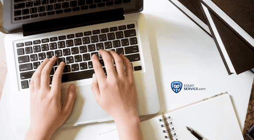 Hands typing on a laptop keyboard with a notebook, pen, and stacked folders on a white desk. The logo "EssayService.com" is visible on the desk.