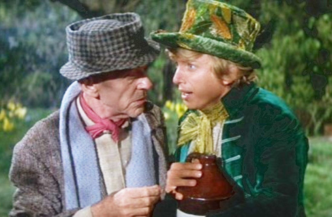 Two men dressed in colorful, old-fashioned costumes talk closely outdoors, one wearing a green hat and jacket, the other in a gray hat, scarf, and red neckerchief.