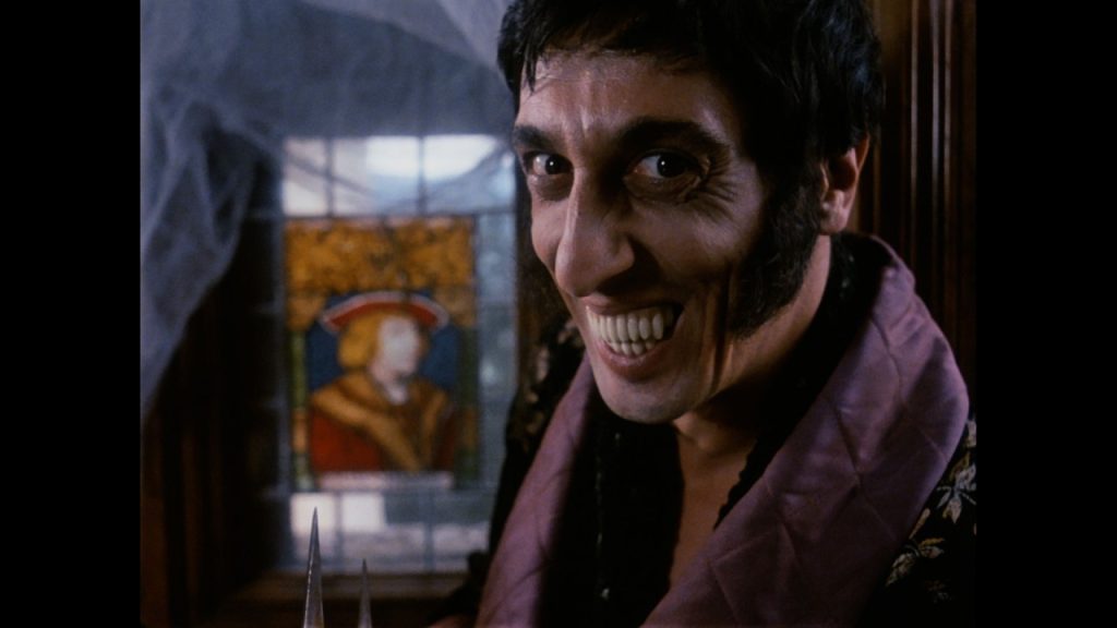 A man with sharp teeth and sideburns grins menacingly in front of a stained glass window depicting a historical figure.