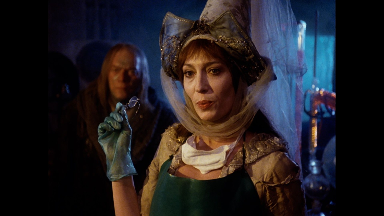 A woman in a medieval-style costume and veil holds up a ring while wearing a glove, with a person standing in the background in a dimly lit room.