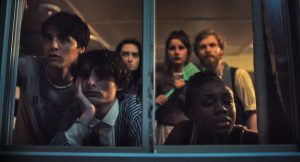 NEON To Release The Comedic Slasher ‘Hell Of A Summer’ On 4K UHD Blu-Ray This July Six people gather in dim lighting, looking intently through a window, with expressions ranging from curiosity to contemplation.