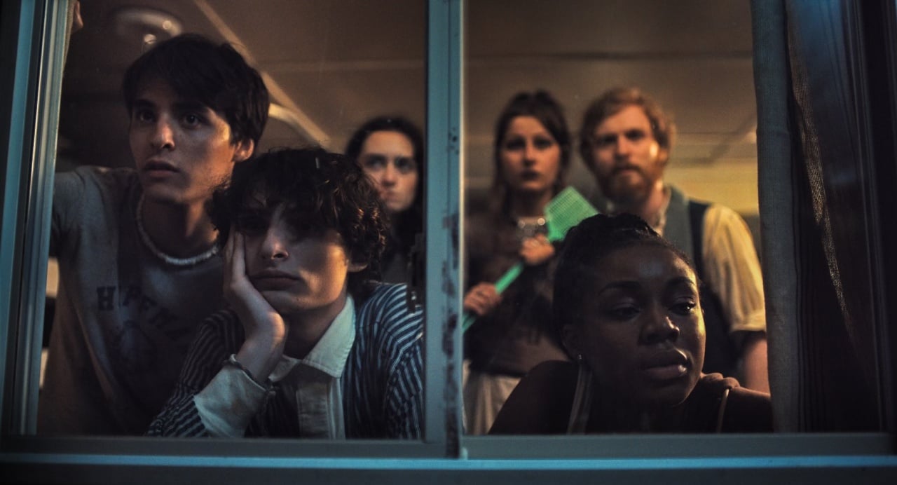 Six people gather in dim lighting, looking intently through a window, with expressions ranging from curiosity to contemplation.