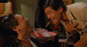 Two men laughing together indoors; one is holding his chest and has a cigar in his mouth, while the other leans back next to a large bowl filled with ice.