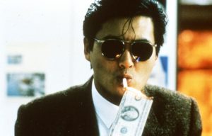 A man in sunglasses and a suit lights a cigarette using a burning banknote.