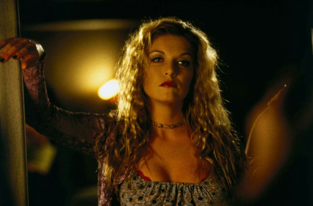 A woman with long, wavy blonde hair and red lipstick stands indoors, leaning against a doorway, looking slightly to the side with a serious expression.