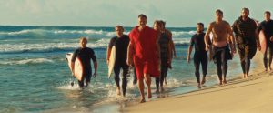 A group of people in wetsuits, some carrying surfboards, walk along a sandy beach near the ocean under clear skies.