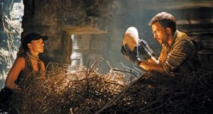 Two people in explorer outfits examine an ancient artifact in a dimly lit, overgrown temple filled with vines and stone carvings.