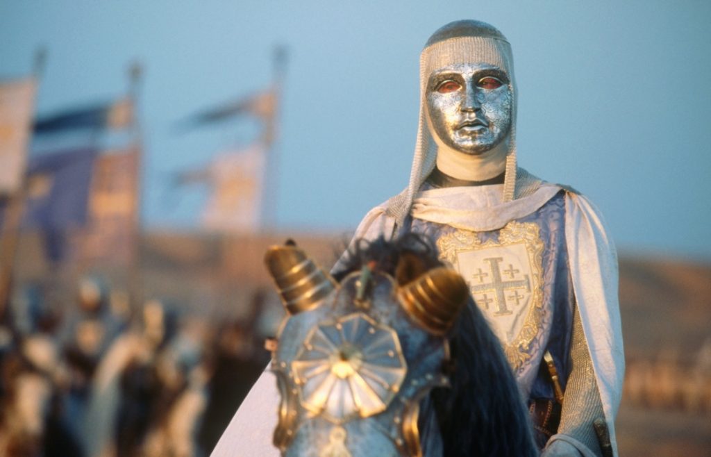 A person in medieval armor with a metallic mask rides a decorated horse. Flags and more armored figures are in the blurred background.