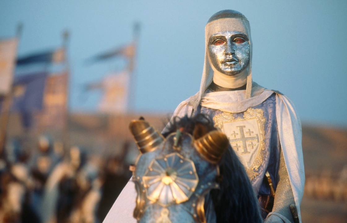 A person in medieval armor with a metallic mask rides a decorated horse. Flags and more armored figures are in the blurred background.
