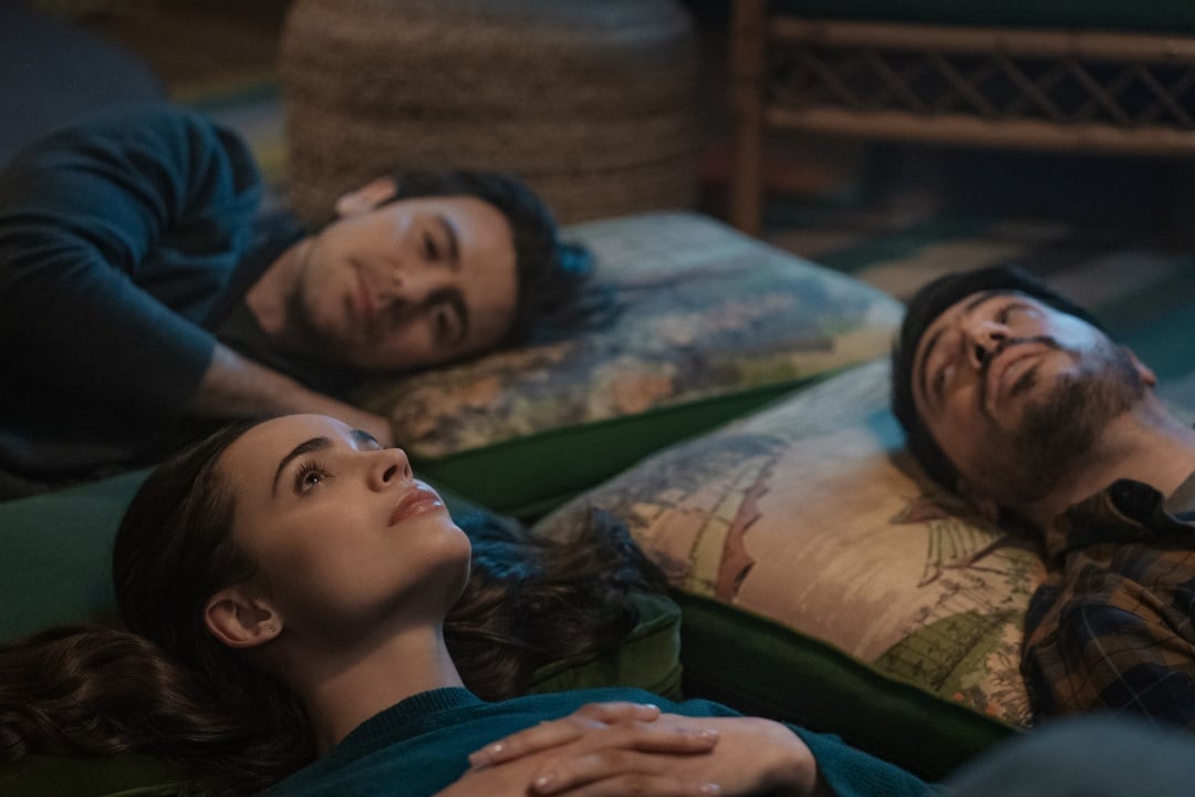 Three people are lying on cushions on the floor, gazing upwards. The room is softly lit, and they appear relaxed and contemplative.