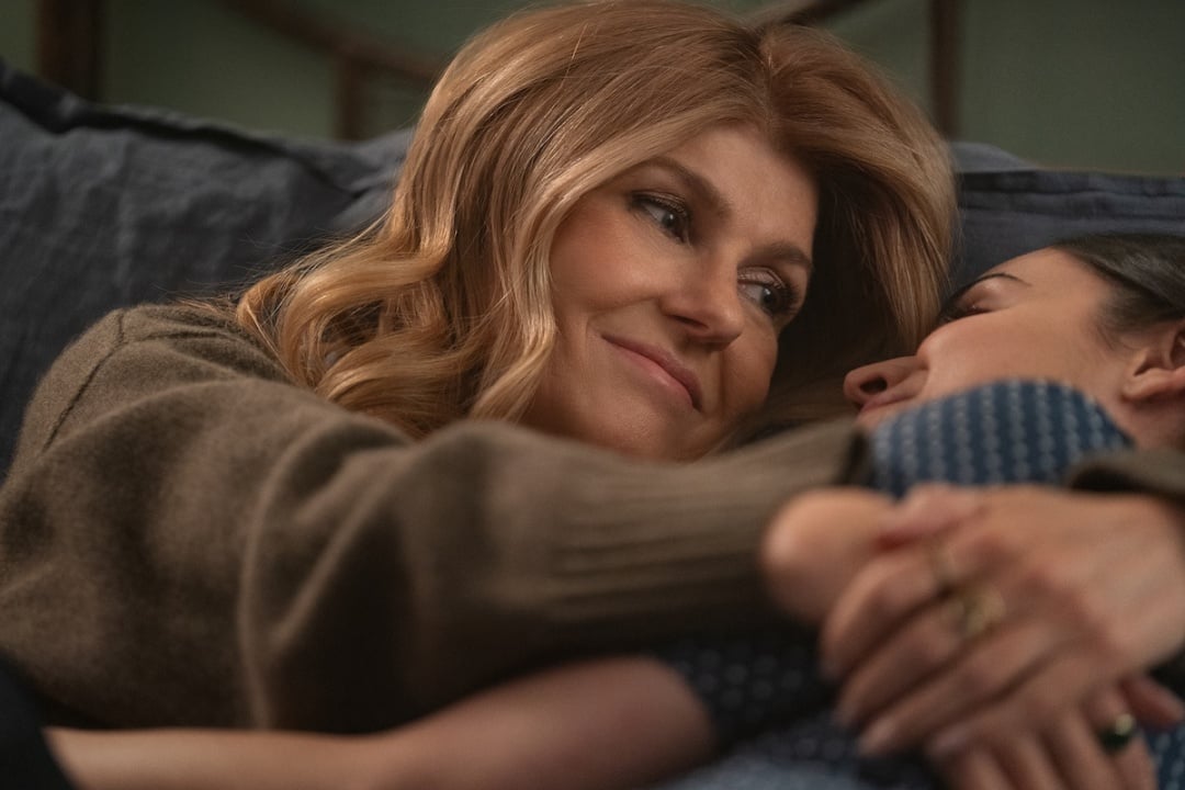 A woman smiles while lying on a couch, embracing another person who is resting on her.