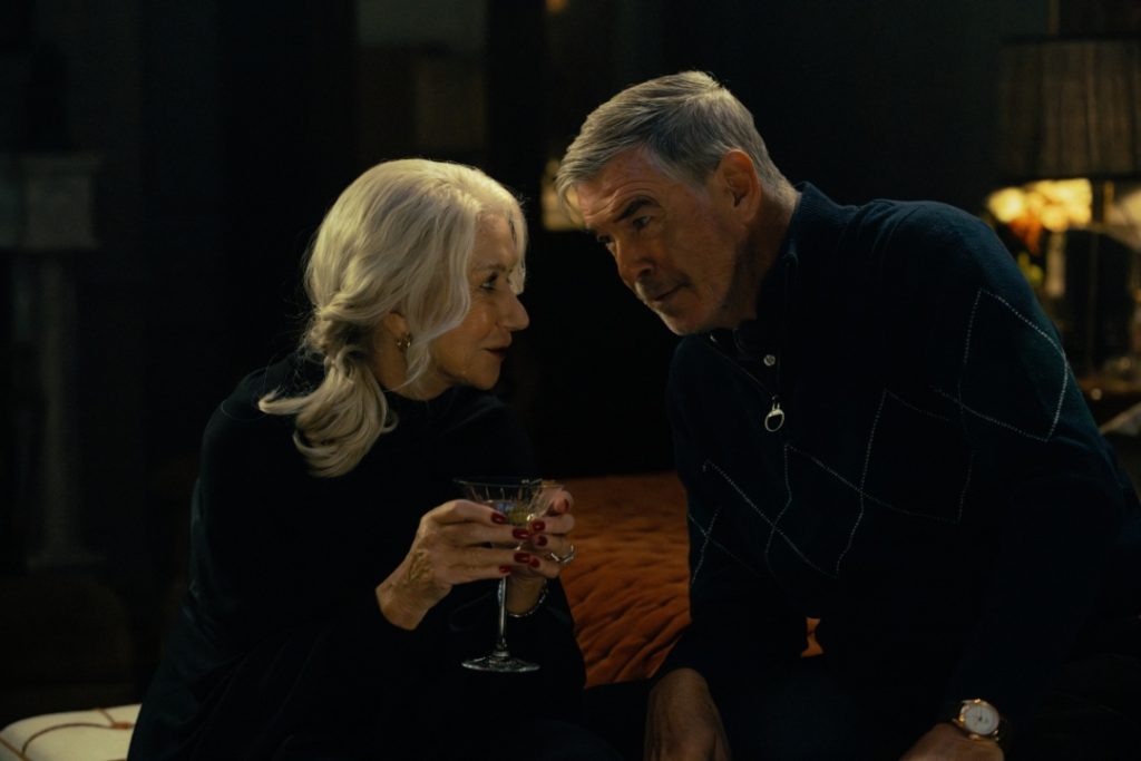 An older woman with white hair holds a glass, sitting close to a man with gray hair, both in dark clothing, in a dimly lit room.