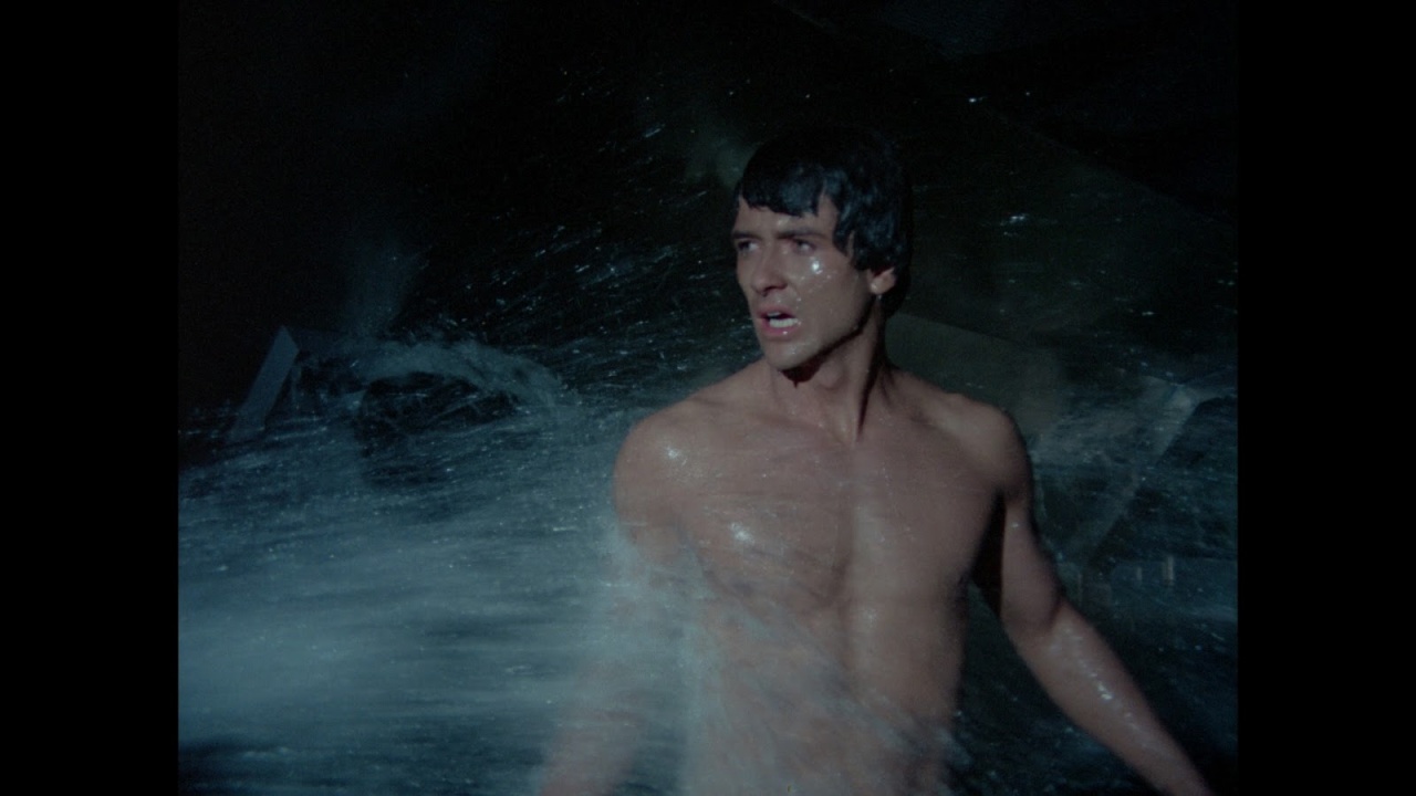 A shirtless man stands waist-deep in water, looking to his side with an intense expression as water splashes around him.