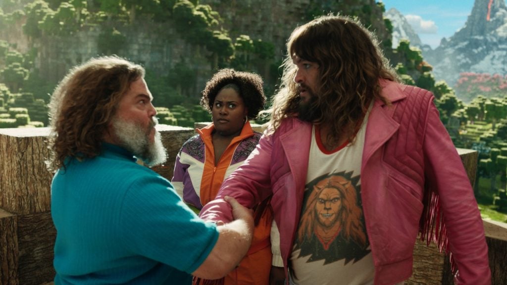 Two bearded individuals, one in a pink jacket and the other in a blue shirt, shake hands outdoors. A person wearing an orange jacket observes them against a scenic mountain background.