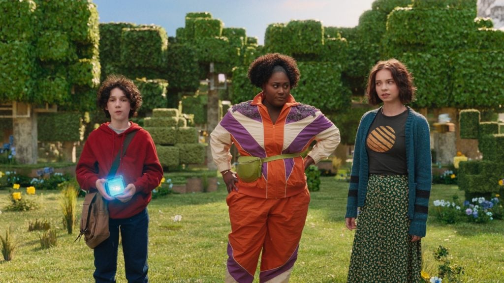 Three people stand in a green park with block-like trees. One holds a glowing cube, another wears a colorful tracksuit, and the third sports a long skirt and patterned shirt.