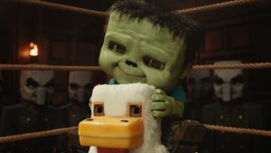 A green cartoon character rides a plush duck on a carousel, surrounded by dark-clad figures with painted faces, inside a dimly lit room.