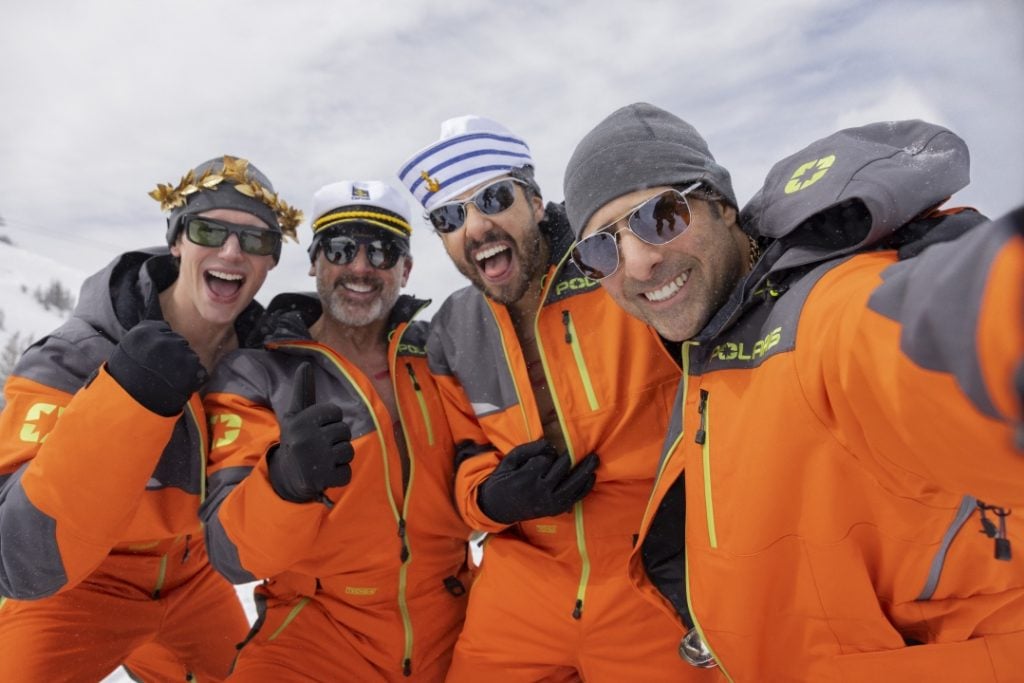 Four people wearing matching orange winter suits and sunglasses pose and smile together outdoors in a snowy setting.