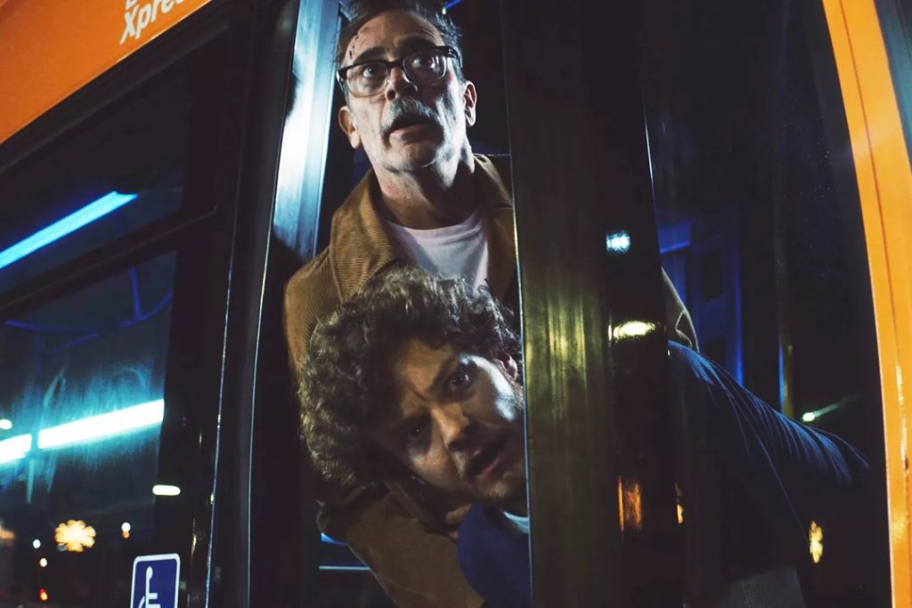 Two men lean out of an open bus door at night, looking in the same direction with concerned expressions.