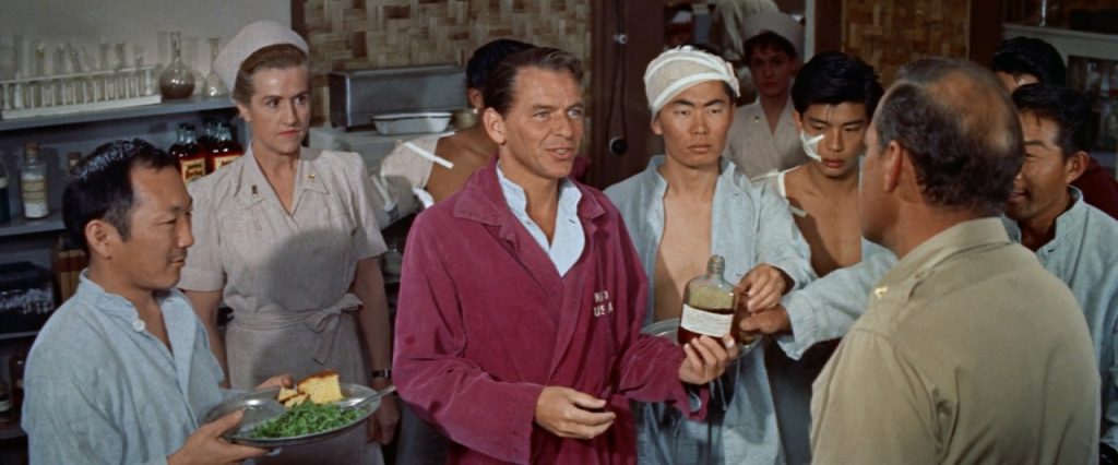A man in a maroon robe holds a bottle while speaking to a uniformed man, surrounded by others in a kitchen or medical setting.