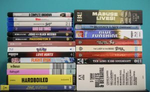 A collection of DVD and Blu-ray cases arranged in two stacks against a solid teal background. The titles vary across genres, including both films and series.