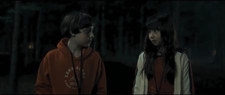 ‘Marshmallow’ Blu-Ray Review – Summer Camp Horror Film Offers Fun Twists & Turns A boy in an orange hoodie and a girl in a jacket stand in a dark, wooded area, looking at each other.