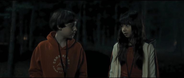 ‘Marshmallow’ Blu-Ray Review – Summer Camp Horror Film Offers Fun Twists & Turns A boy in an orange hoodie and a girl in a jacket stand in a dark, wooded area, looking at each other.