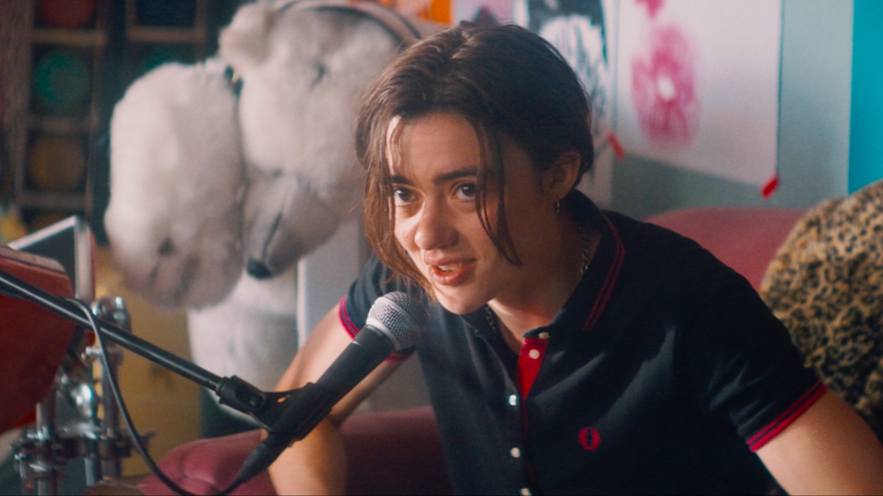 A person with short brown hair speaks into a microphone while sitting on a red chair in a colorful room with posters and a large stuffed animal in the background.