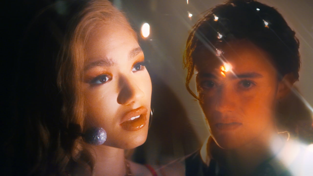 Two people are shown in soft, dramatic lighting with lens flares; one has blonde hair and wears a headset, while the other has short dark hair and a serious expression.