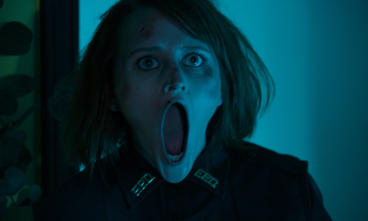 Person in a dark uniform with wide eyes and an elongated open mouth, standing in a dimly lit room.