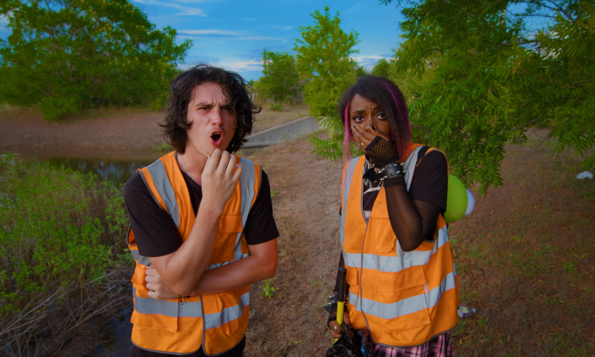 Two people wearing orange safety vests stand outdoors. The person on the left has a surprised expression with a hand on their chin, while the person on the right covers their mouth, holding a camera.