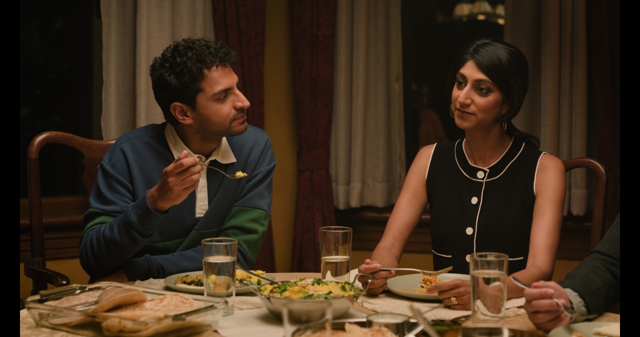 Two people sitting at a dining table, sharing a meal. The man is holding a fork with food, and they are looking at each other. Several dishes and glasses of water are on the table.