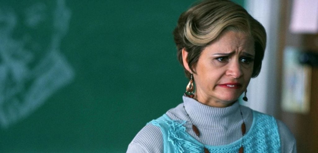 A woman with short, styled hair and a concerned expression stands in front of a green chalkboard, wearing a light blue vest over a gray turtleneck.