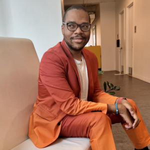 A person wearing an orange suit and glasses sits on a beige bench in a modern hallway, looking to the side and smiling slightly.