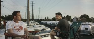 Two men sit in camping chairs on top of an RV, surrounded by a lot full of similar RVs, with power lines and houses in the background.