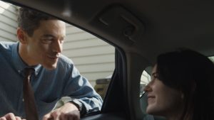 A man leans on a car door, smiling at a woman inside. Both are having a conversation, with a house in the background.