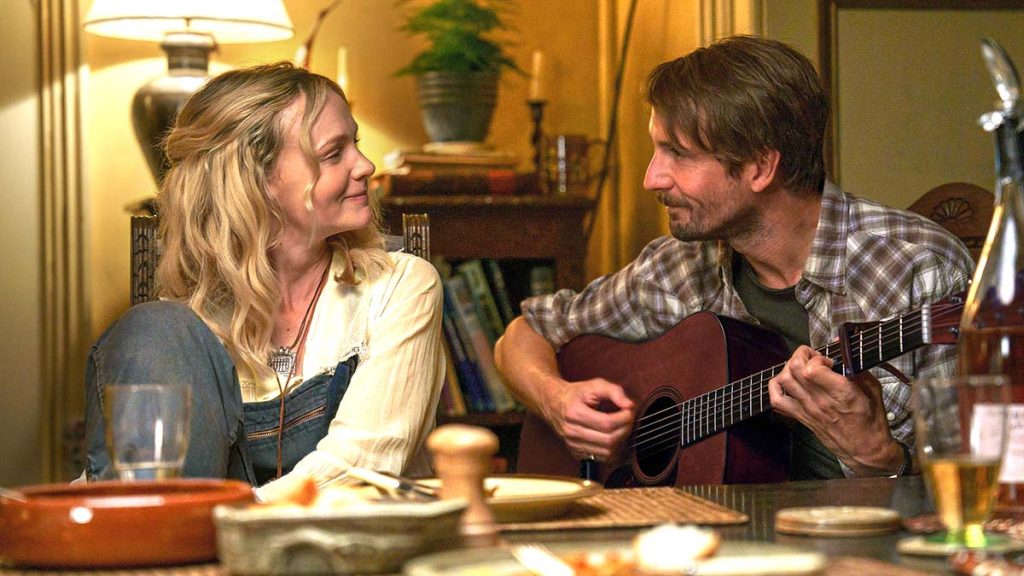 A woman smiles at a man playing an acoustic guitar at a dining table, with food and drinks in the foreground and warm lighting in the background.