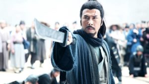 A man with a scar on his forehead points a sword forward. He wears traditional clothing. There is a crowd in the background.