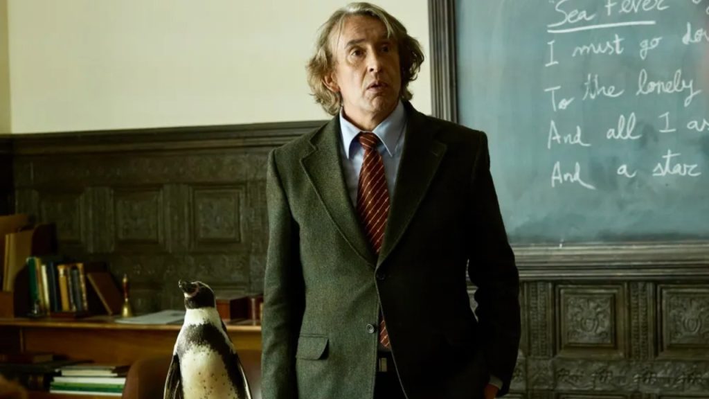 A man in a suit stands next to a penguin in a classroom with a chalkboard in the background.