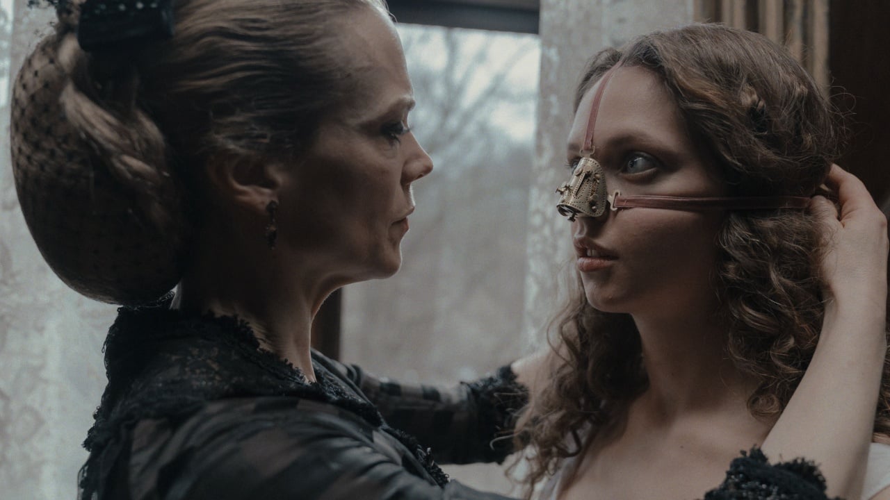 A woman adjusts a steampunk-like mask on another woman's face in a dimly lit room.