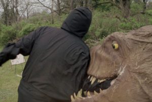 A person in a black hooded outfit is being bitten on the arm by a large, artificial dinosaur head outdoors.