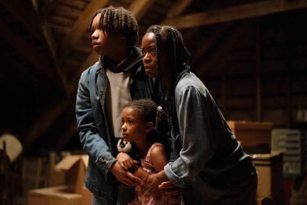 Three people, an adult and two children, huddle together in a dimly lit attic, surrounded by boxes. One child holds a stuffed panda.