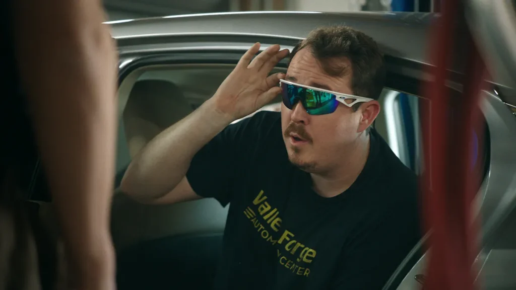 Man wearing sunglasses sits in a car, adjusting them with his hand. He's wearing a T-shirt with "Valle Forge Auto Center" printed on it.