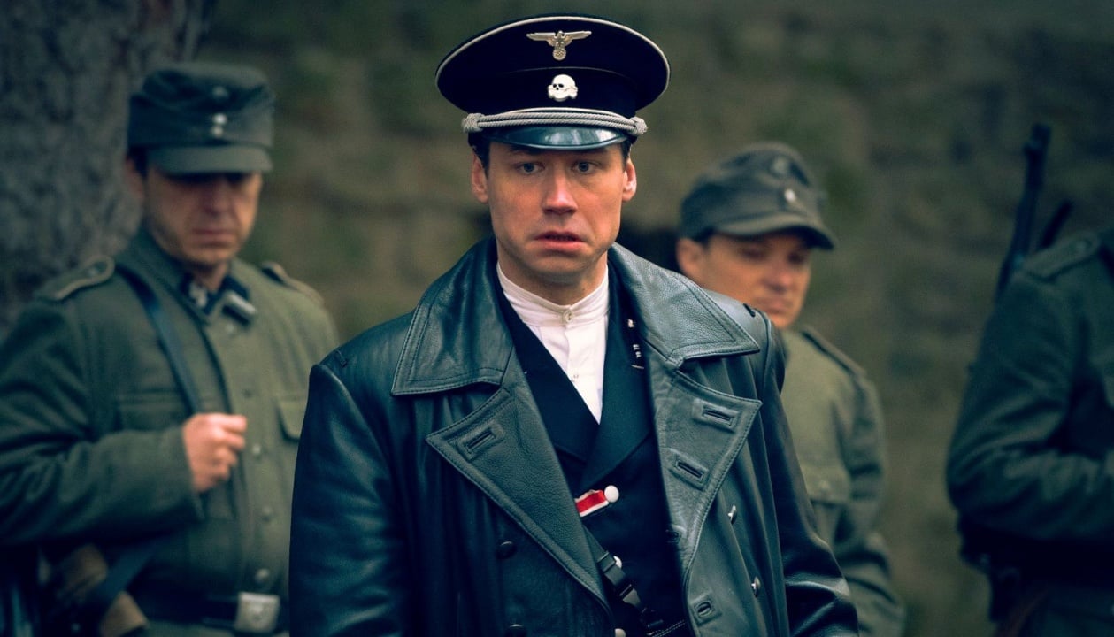 A man in a black military uniform and coat stands outdoors, accompanied by others in similar attire.