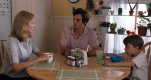 Three people sit at a kitchen table with mugs, a bowl, and medicine bottles; potted plants are visible on a shelf by the window in the background.
