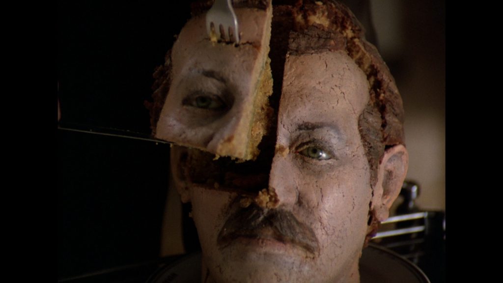 A realistic cake shaped like a man's head is being sliced, with a piece containing an eye being lifted by a fork.