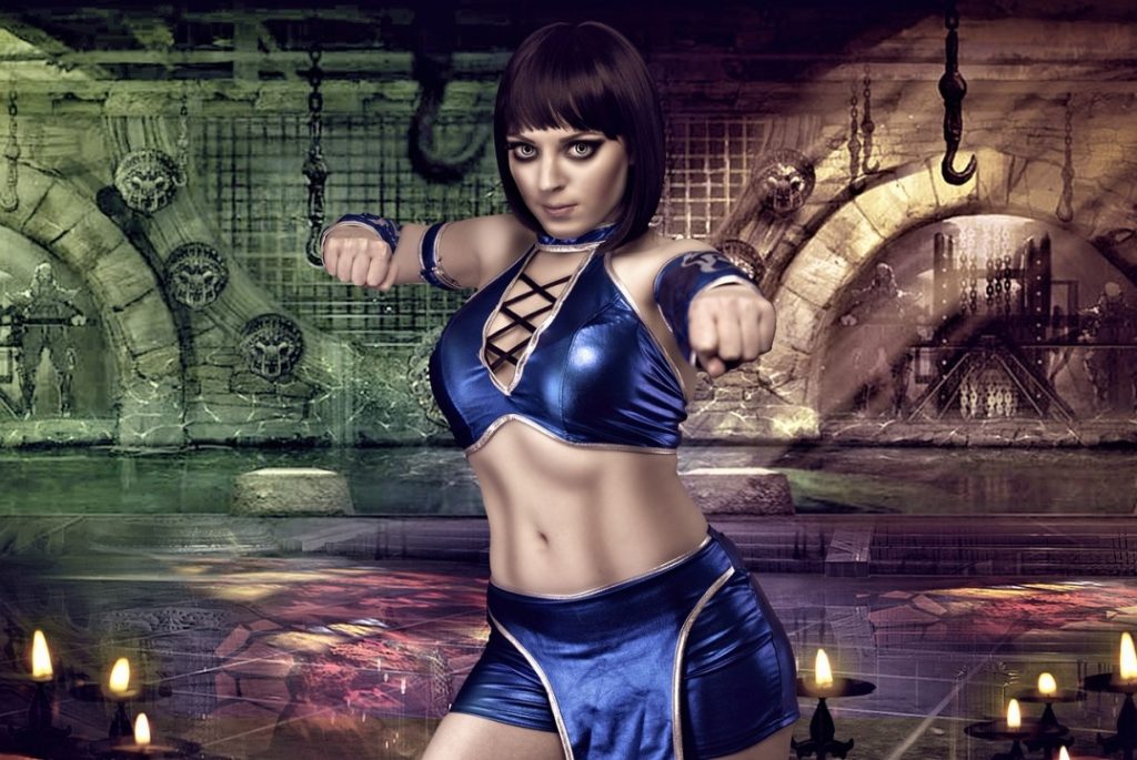 A woman in a blue warrior costume poses with fists raised in an underground setting with stone walls and chains.